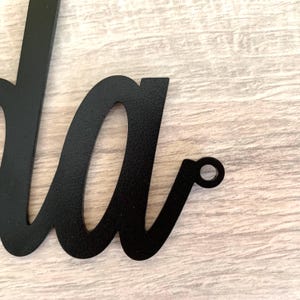 May include: A black metal wall sign featuring a cursive script font. The visible portion of the sign shows the lowercase letters 'da' with a small circular mounting hole integrated into the end of the final letter's flourish for hanging.