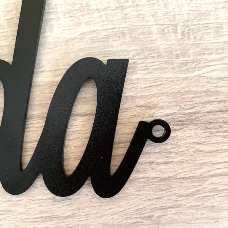 May include: A black metal wall sign featuring a cursive script font. The visible portion of the sign shows the lowercase letters 'da' with a small circular mounting hole integrated into the end of the final letter's flourish for hanging.