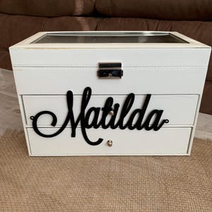 May include: A white rectangular jewelry box with a clear glass lid and a silver metal latch. The front features two drawers with small silver knobs, and the name Matilda is written in large black cursive script across the top drawer front.