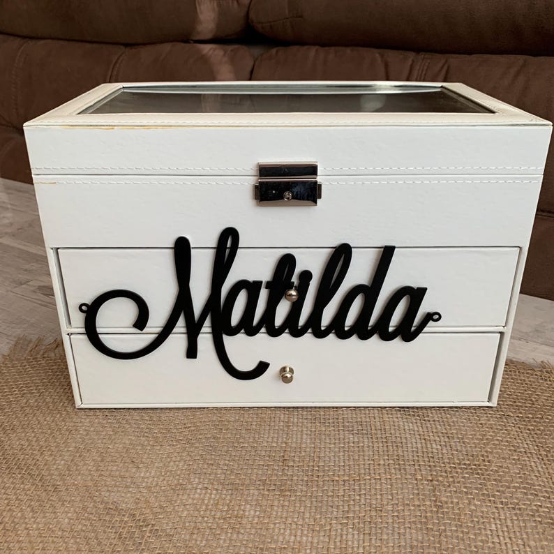 May include: A white rectangular jewelry box with a clear glass lid and a silver metal latch. The front features two drawers with small silver knobs, and the name Matilda is written in large black cursive script across the top drawer front.