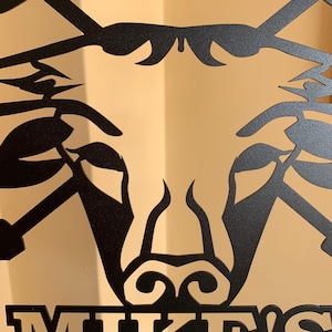 May include: Black metal wall art featuring a stylized bull's head design. The artwork has a matte finish and includes the name "MIKE'S" at the bottom. The bull's head is depicted with horns and a detailed facial structure, suitable for home decor.