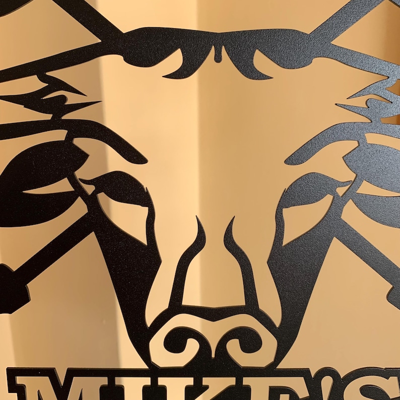 May include: Black metal wall art featuring a stylized bull's head design. The artwork has a matte finish and includes the name "MIKE'S" at the bottom. The bull's head is depicted with horns and a detailed facial structure, suitable for home decor.