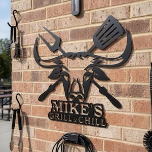 May include: Black metal wall art featuring a barbecue theme. The design includes a spatula, fork, and the text "MIKE'S GRILL & CHILL". Additional grilling tools and a thermometer are also visible on the brick wall.
