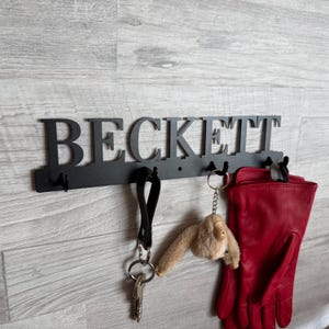 Könnte beinhalten: Schwarzer Metall-Wand-Schlüsselhalter mit dem ausgeschnittenen Namen "BECKETT". Der Schlüsselhalter hat mehrere Haken mit einem Schlüsselbund, einem Plüschtier und einem Paar roter Handschuhe.