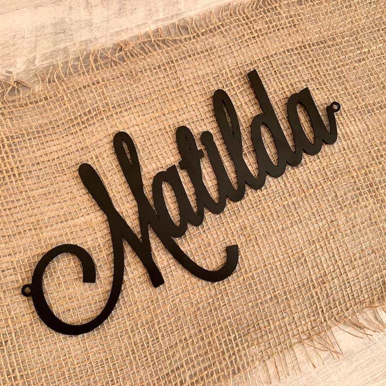 May include: A black, script-style wall sign featuring the name Matilda in a continuous, flowing cursive font. The sign is constructed from a flat, solid material and includes small circular mounting holes at both the far left and far right ends.