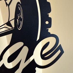 Op de afbeelding: Zwart metalen bord met een silhouet van een auto en het woord "Garage" in een gestileerd lettertype.