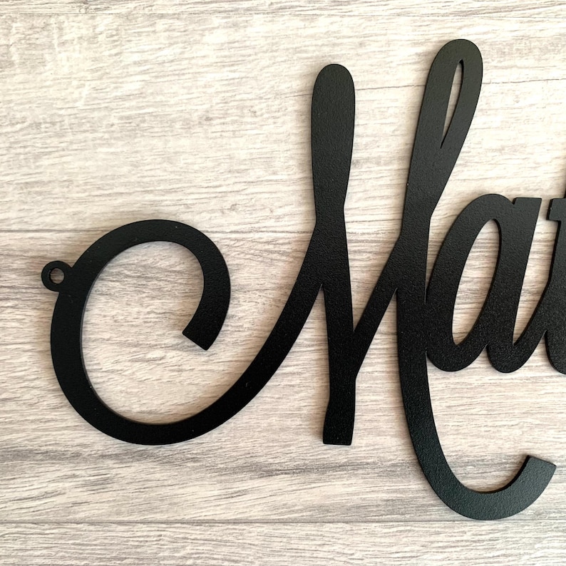 May include: A matte black metal wall sign featuring the cursive letter M. The script font has a smooth finish and a small circular mounting hole on the far left side of the letter. The design is a single continuous piece of cut metal.