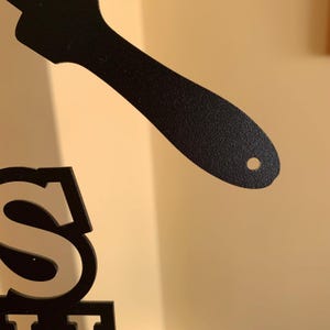May include: Black metal decorative sign with the letter "S" and a spatula-shaped design. The sign is made of a textured metal and is against a neutral background.