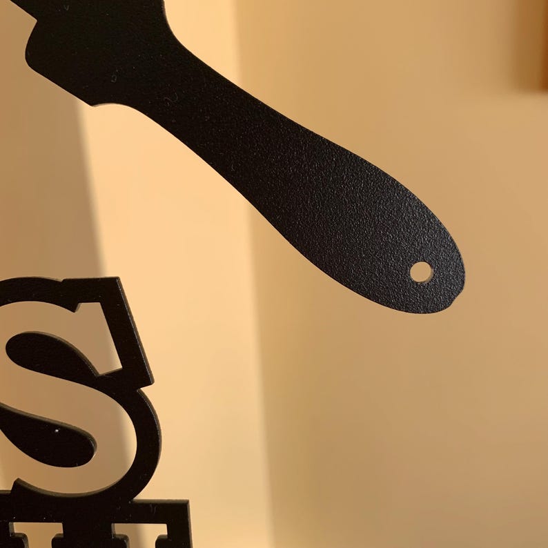 May include: Black metal decorative sign with the letter "S" and a spatula-shaped design. The sign is made of a textured metal and is against a neutral background.