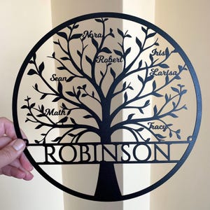 以下が含まれることがあります： 「ROBINSON」という名字が書かれた黒い金属製のファミリーツリーの壁飾り。枝には「Nora」、「Robert」、「Sean」、「Math」、「Iris」、「Larisa」、「Tracy」という家族の名前が書かれている。