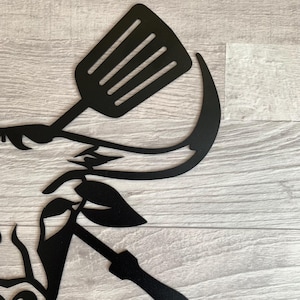 May include: Black metal wall art featuring a spatula, a curved blade, and leaf accents. The design is cut from metal and is suitable for kitchen or outdoor decor. The artwork is mounted on a light wood-grain surface.