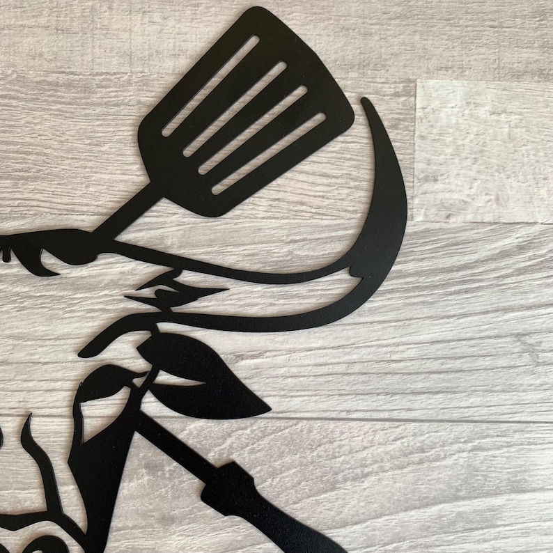 May include: Black metal wall art featuring a spatula, a curved blade, and leaf accents. The design is cut from metal and is suitable for kitchen or outdoor decor. The artwork is mounted on a light wood-grain surface.