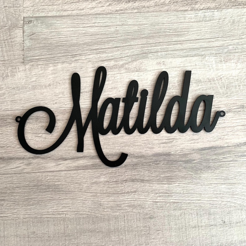 May include: A black wall sign featuring the name Matilda written in a continuous cursive script font. The letters are connected in a flowing style, and the sign includes small circular mounting holes at the far left and far right ends.