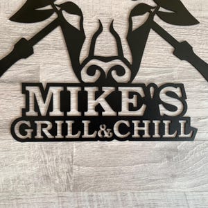 May include: Black metal sign with the text "MIKE'S GRILL & CHILL" in a bold, stylized font. The sign features decorative elements above the text, possibly for outdoor use. The sign is mounted on a light gray wooden surface.