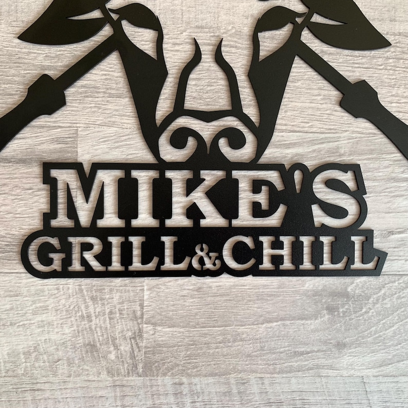 May include: Black metal sign with the text "MIKE'S GRILL & CHILL" in a bold, stylized font. The sign features decorative elements above the text, possibly for outdoor use. The sign is mounted on a light gray wooden surface.