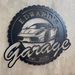 Op de afbeelding: Zwart metalen bord met een bandenprofiel, met een silhouet van een sportwagen en de tekst "Edward's Garage est. 2025".