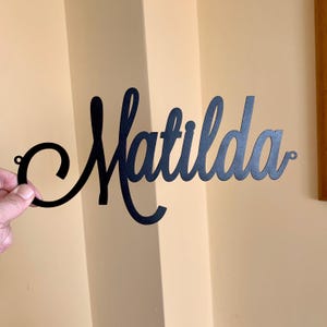 May include: A matte black metal wall sign featuring the name Matilda written in a continuous cursive script font. The design includes small circular mounting holes at both the far left and far right ends of the lettering for wall attachment.