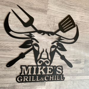 May include: Black metal wall art featuring a barbecue theme. The design includes a pitchfork, spatula, and other grilling tools, along with the text "MIKE'S GRILL & CHILL". The artwork is mounted on a wooden surface.