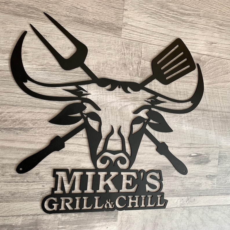May include: Black metal wall art featuring a barbecue theme. The design includes a pitchfork, spatula, and other grilling tools, along with the text "MIKE'S GRILL & CHILL". The artwork is mounted on a wooden surface.
