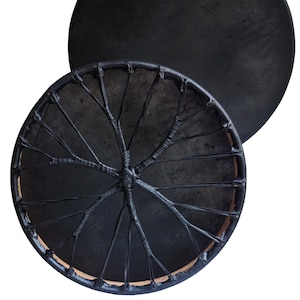 May include: Two black, round drums. The front drum has a dark, textured surface with a web-like design in the center. The second drum is in the background, and has a smooth, dark surface. The drums are likely used for music or spiritual practices.