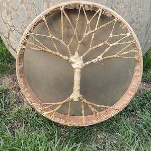 May include: A round, wooden frame drum with a natural leather head. The drum is decorated with a woven tree of life design in a light brown color.
