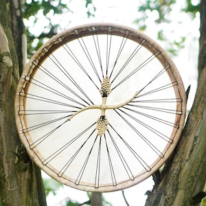May include: A round, handmade drum with a natural wood frame and a stretched, off-white drumhead. Black cords radiate from the center, and the drum is suspended in a tree, creating a rustic, natural aesthetic.