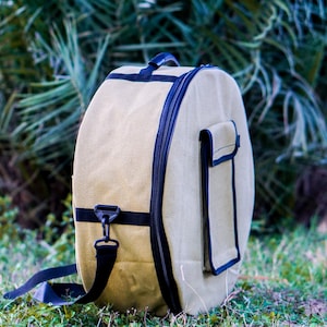 May include: A beige canvas drum bag with a black zipper and strap. The bag has a black pocket on the side.