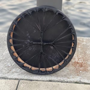 May include: A black hand drum with a wooden frame and a black leather head. The drum is strung with black leather lacing.