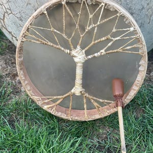 May include: A brown leather drum with a tree of life design and a brown leather mallet. The drum is made of wood and has a natural finish. The mallet has a wooden handle and a leather head.