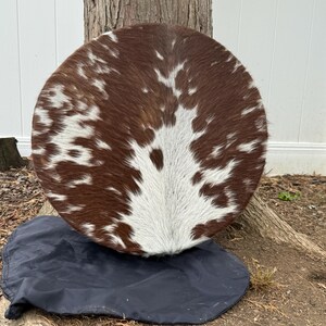 May include: A round, brown and white cowhide rug. The rug has a natural pattern and texture, with a mix of brown and white patches. It is placed on a black fabric.