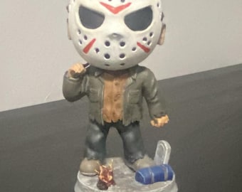 May include: A bobblehead figure featuring a character wearing a white hockey mask with red accents, a brown shirt, and a dark green jacket. The figure stands on a circular base with a small campfire, a blue sleeping bag, and a tombstone accessory.