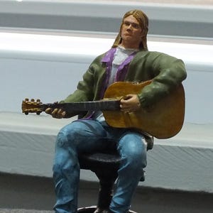 Op de afbeelding: Een miniatuurbeeldje van een persoon die een akoestische gitaar bespeelt. De figuur zit op een zwarte stoel, draagt een blauwe spijkerbroek, een groene jas en een wit overhemd met een paarse kraag. De gitaar is bruin en geel.