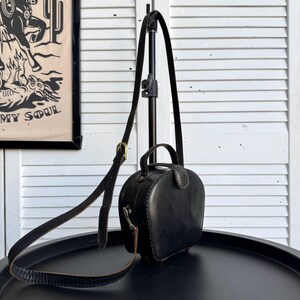 May include: A small, black leather handbag with a rounded shape and a top handle. It has a long, adjustable shoulder strap and a small buckle. The bag is on a black surface.