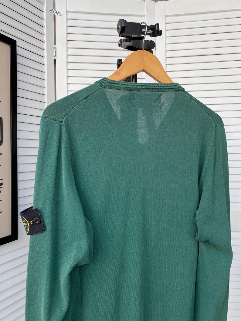Pu&ograve; includere: Un maglione verde a maniche lunghe con una piccola toppa circolare sul braccio sinistro. La toppa ha un design a bussola con sfondo giallo e dettagli neri.