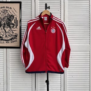 Könnte beinhalten: Rote Adidas-Trainingsjacke mit weißen Akzenten und dem Bayern München-Logo. Die Jacke hat einen durchgehenden Reißverschluss, weiße Streifen auf den Ärmeln und einen marineblauen Saum. Das Adidas-Logo befindet sich auf der Brust.