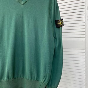 Pu&ograve; includere: Un maglione verde a collo a V con una toppa Stone Island nera e gialla sulla manica destra.
