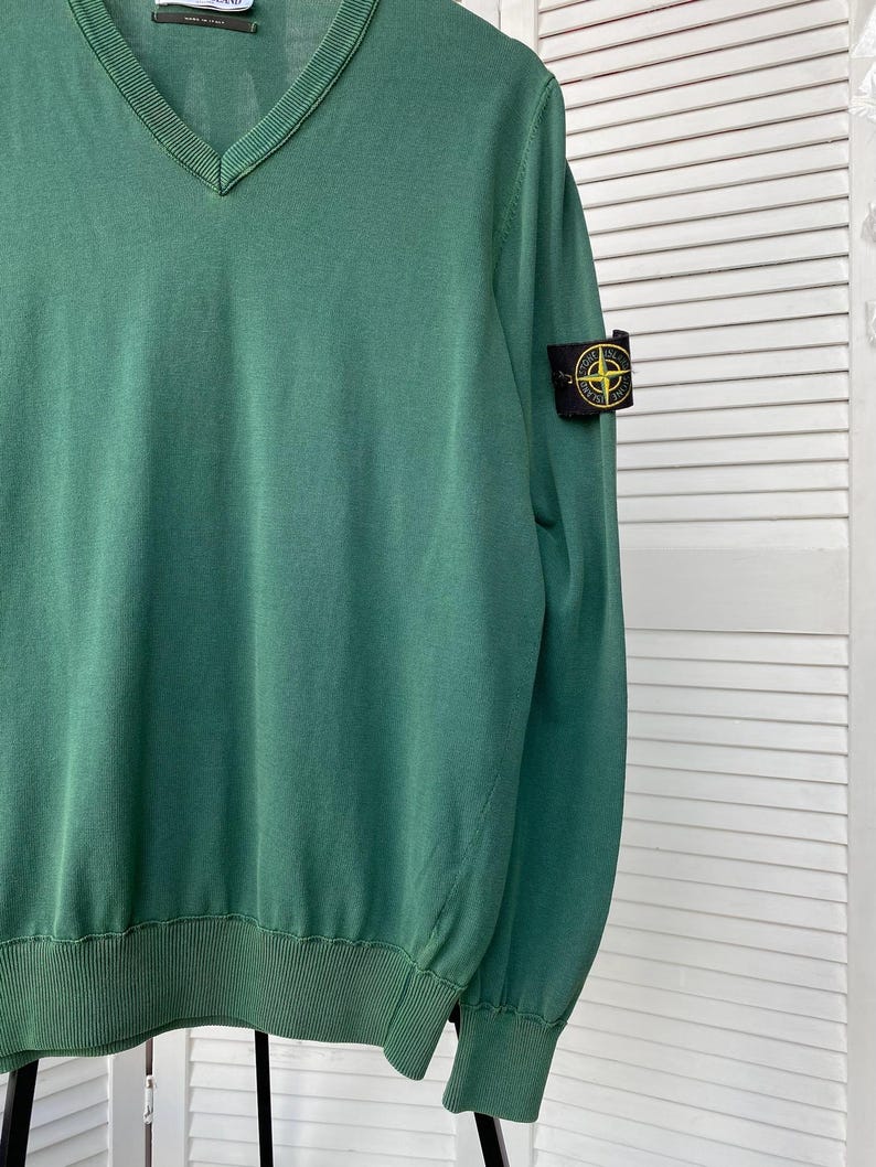 Pu&ograve; includere: Un maglione verde a collo a V con una toppa Stone Island nera e gialla sulla manica destra.