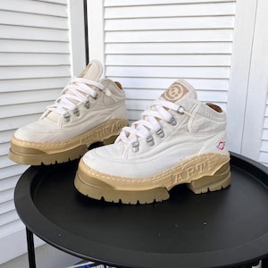 Puede incluir: Un par de zapatillas blancas de lona de caña alta con una suela beige gruesa. Las zapatillas tienen un logotipo en el lateral que dice "The Art" y "Art" en el otro lado.