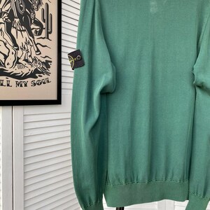 Pu&ograve; includere: Un maglione verde a maniche lunghe con una piccola toppa nera e gialla sulla manica sinistra. La toppa ha un design a bussola.