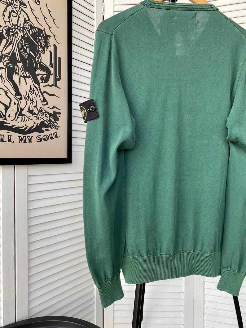 Pu&ograve; includere: Un maglione verde a maniche lunghe con una piccola toppa nera e gialla sulla manica sinistra. La toppa ha un design a bussola.