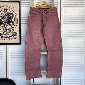 Könnte beinhalten: Eine Jeans aus verblassten rosa Denim mit einem rohen Saum. Die Jeans hat eine lockere Passform und hängt an einem Holzbügel.