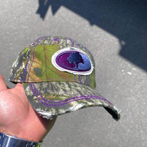May include: A camouflage baseball cap with a purple and blue embroidered patch featuring a tree silhouette. The cap has a purple stitch around the brim.