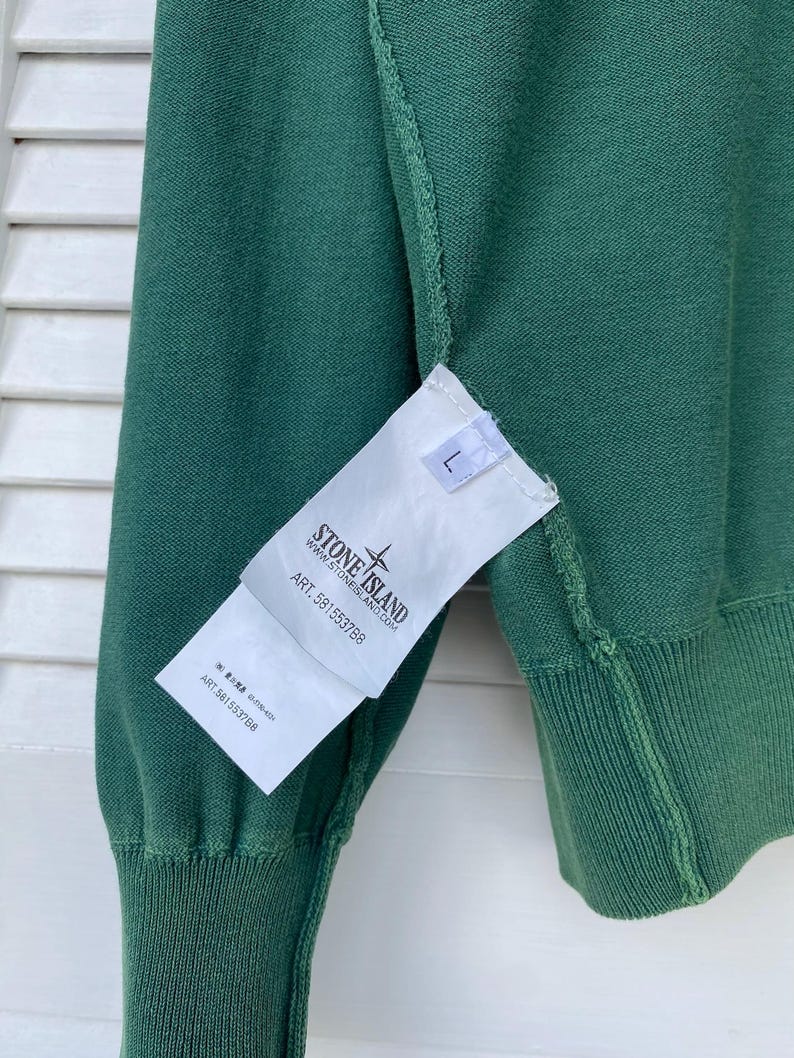 Pu&ograve; includere: Un maglione verde Stone Island con un'etichetta bianca che dice "Stone Island" e "Art. 581553788".