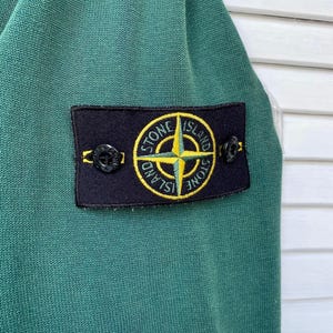 Pu&ograve; includere: Una camicia verde a maniche lunghe con una toppa nera che presenta una bussola gialla e il testo "Stone Island".