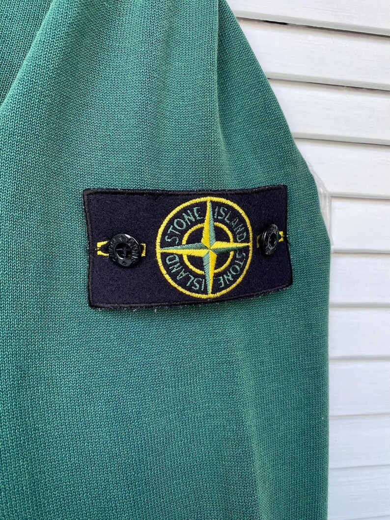 Pu&ograve; includere: Una camicia verde a maniche lunghe con una toppa nera che presenta una bussola gialla e il testo "Stone Island".