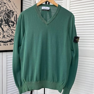 Pu&ograve; includere: Un maglione verde a collo a V con maniche lunghe e una patch con il logo Stone Island sulla manica sinistra. Il maglione &egrave; appeso a un appendiabiti in legno.