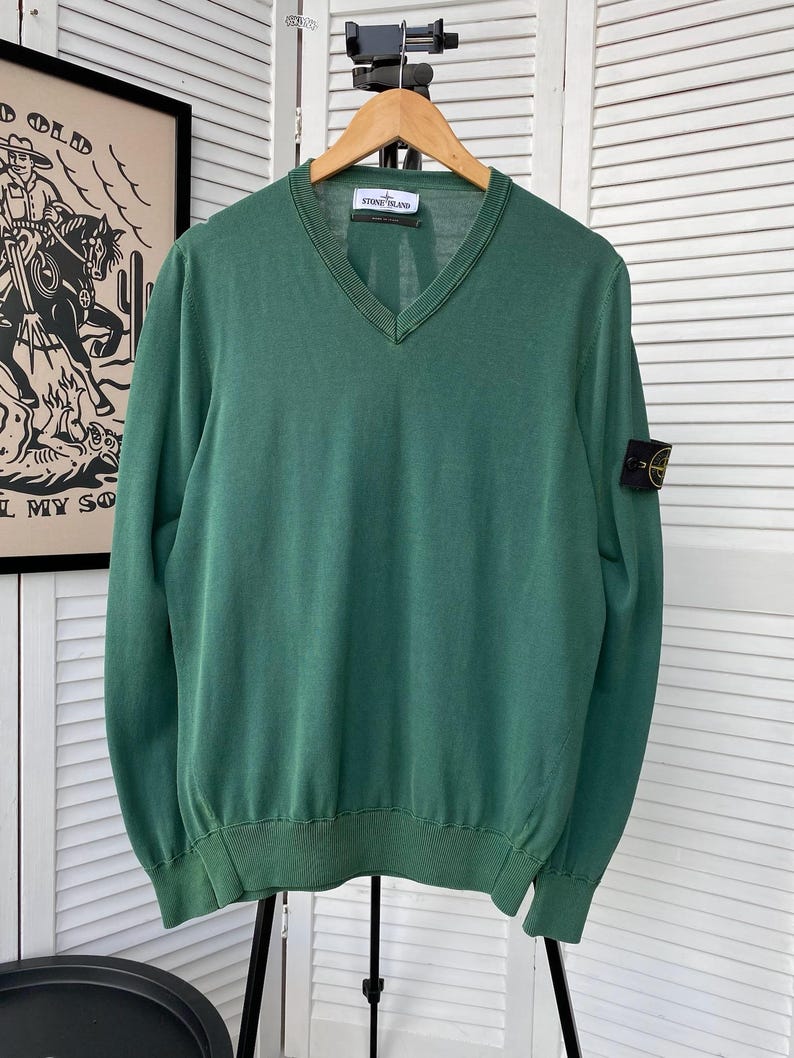 Pu&ograve; includere: Un maglione verde a collo a V con maniche lunghe e una patch con il logo Stone Island sulla manica sinistra. Il maglione &egrave; appeso a un appendiabiti in legno.