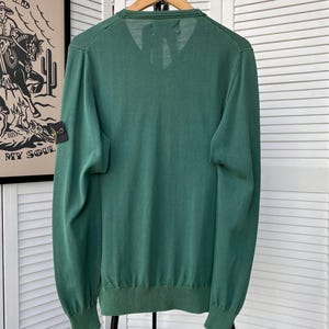 Pu&ograve; includere: Un maglione verde a maniche lunghe con scollo a V e una piccola toppa nera sul braccio sinistro. La toppa ha un design a bussola e il testo "Stone Island".