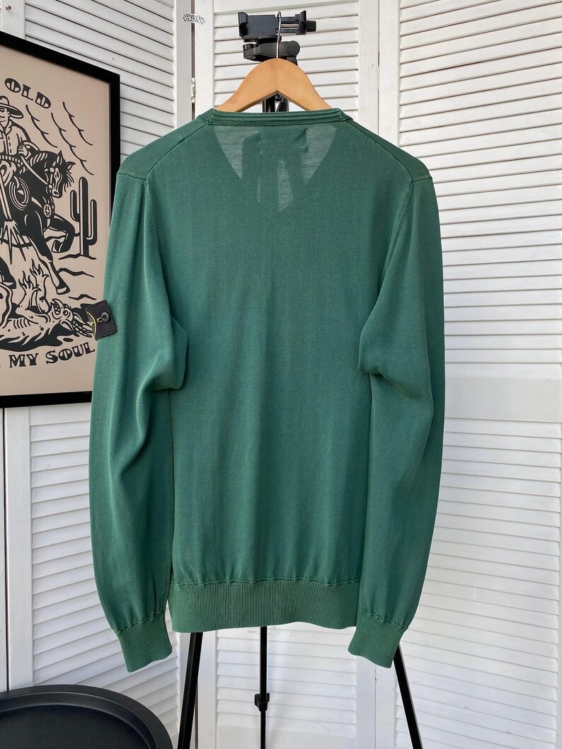 Pu&ograve; includere: Un maglione verde a maniche lunghe con scollo a V e una piccola toppa nera sul braccio sinistro. La toppa ha un design a bussola e il testo "Stone Island".