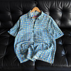 May include: A short-sleeved button-down shirt with a blue background and a repeating pattern of small squares in yellow, green, and brown. The shirt is hanging on a wooden hanger against a black leather couch.